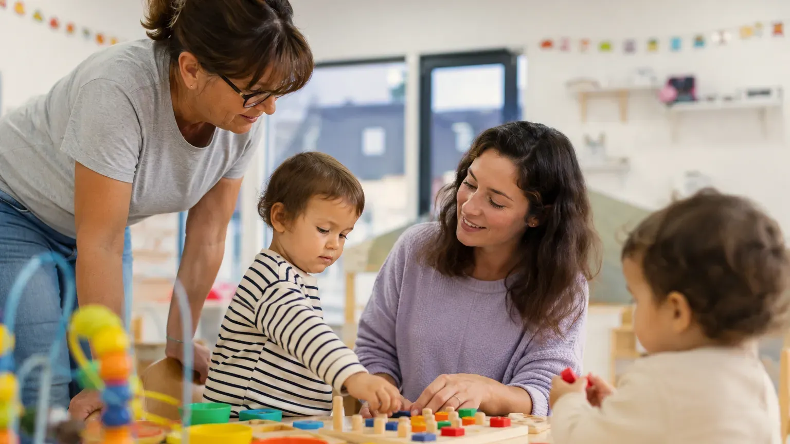 Adulte en reconversion professionnelle participant à une activité d'éveil avec de jeunes enfants dans une crèche, sous la supervision d'une professionnelle de la petite enfance.