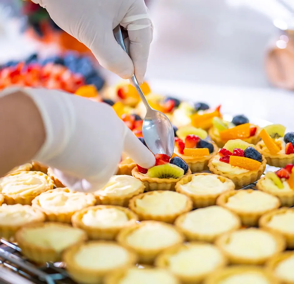 Apprenant en formation pâtisserie réalisant des tartelettes aux fruits dans un atelier pratique, en déposant des morceaux de fraise sur une crème pâtissière